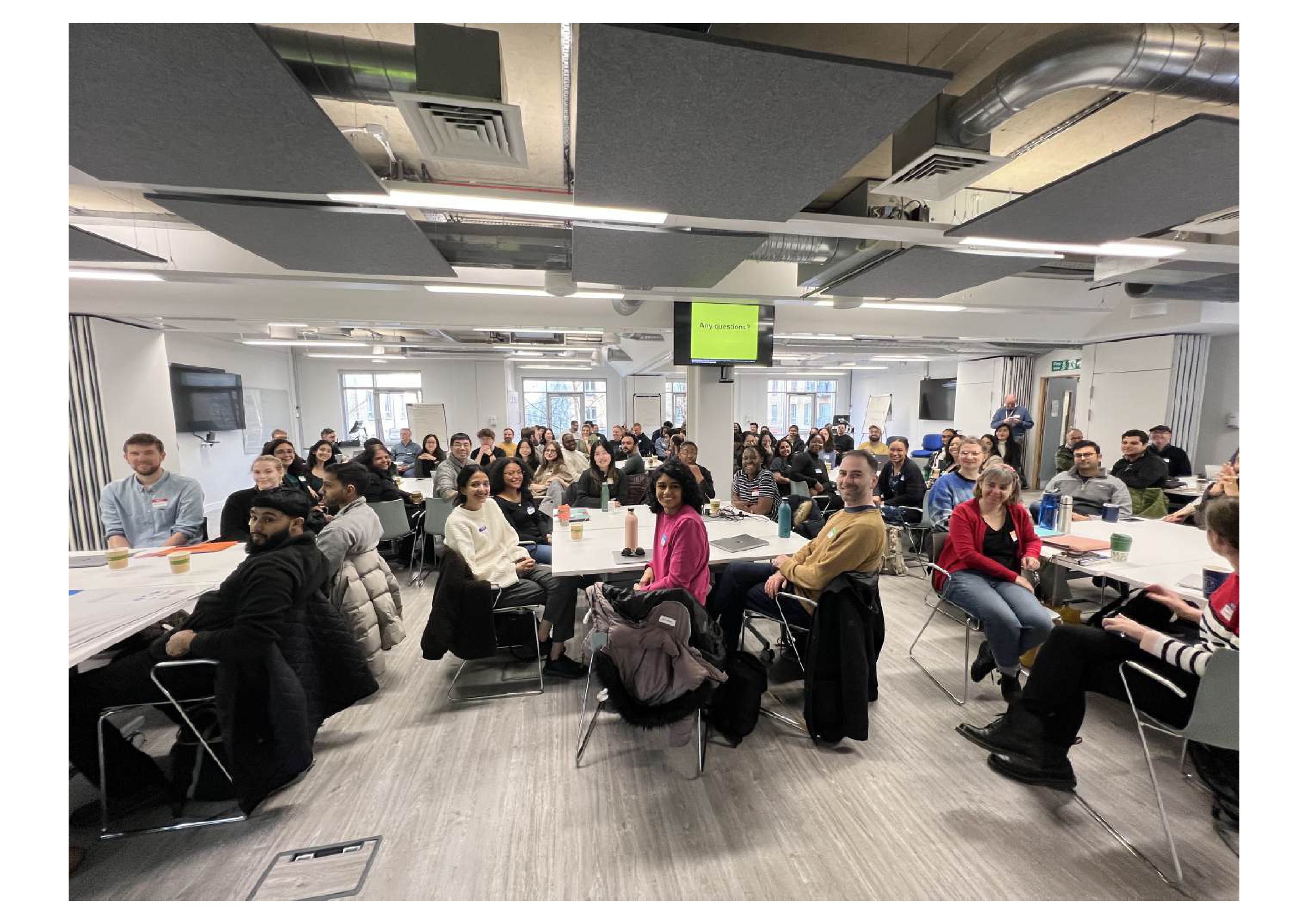 Hackday participants sitting in groups around tables looking towards the camera